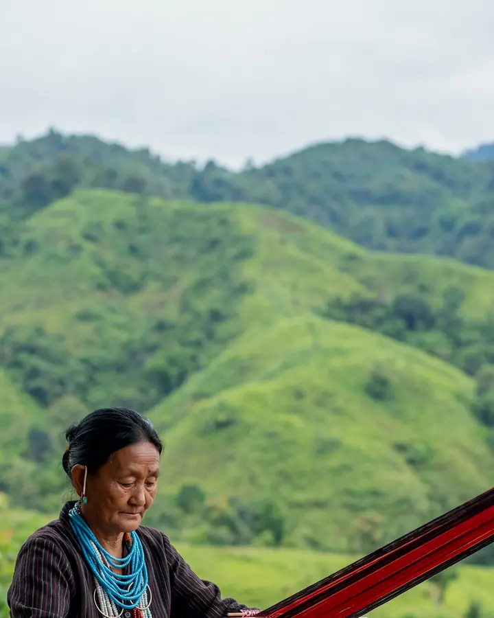 An Adi woman in traditional dress weaving on a backstrap loom on a veranda, with baskets of oranges beside her.