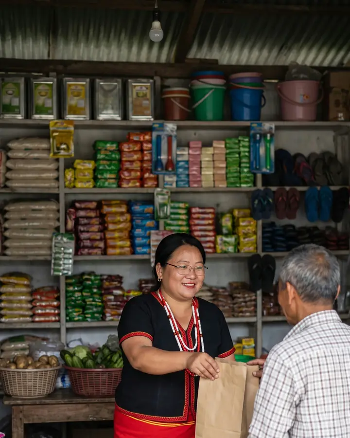 A smiling Adi woman shopkeeper in glasses, black Adi blouse and bead necklaces, hands a paper bag to an older customer across the counter of a busy, well-stocked village shop.